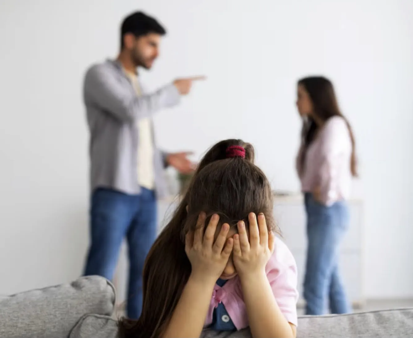 Una bambina si copre il viso con le mani mentre sullo sfondo, sfocati, due genitori discutono